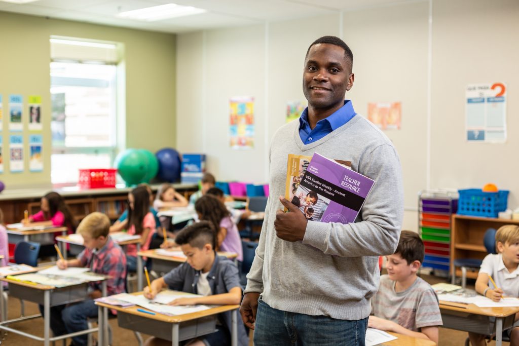 2018 Studies Weekly Photoshoot | Male teacher standing holding book, workbook.