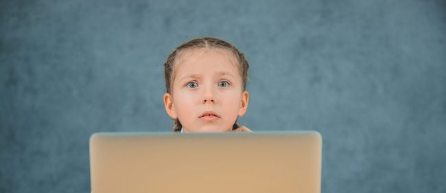 Confused schoolgirl with braids sitting in front of grey laptop isolated on grey background.