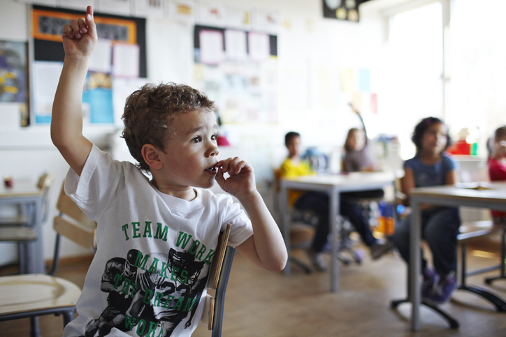 Cute boy with raised hand in classroom Have small class size for summer school program for elementary studentss