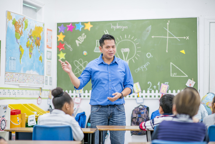 Male Teacher Leading The Class Teacher in front of classroom.