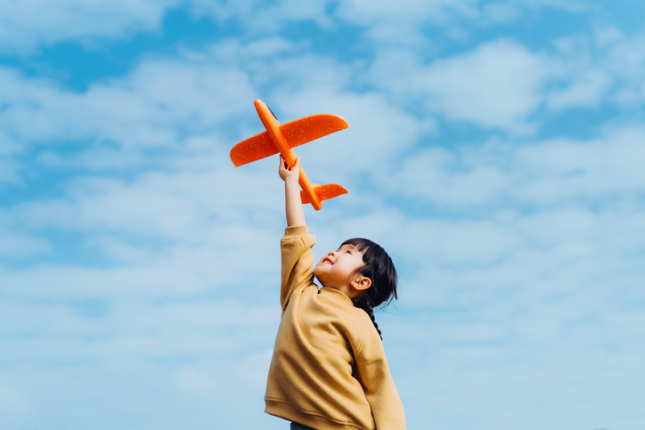 Happy lovely little Asian girl having fun outdoors, playing with airplane toy and smiling joyfully in park on a lovely sunny day against beautiful blue sky Learning Intentions child is aiming for with a plane