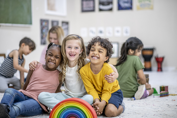 A small group of three school aged children huddle together on an area rug in their classroom as they pose for a portrait. They are each dressed casually and rainbow blocks can be seen stacked in front of them.
