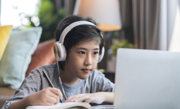 Asaian child Boy kid taking an E-learning learning online class from school teacher by digital remote device laptop internet meeting in living room ,asia teen wear headphone using laptop e learning class at home Student taking online test