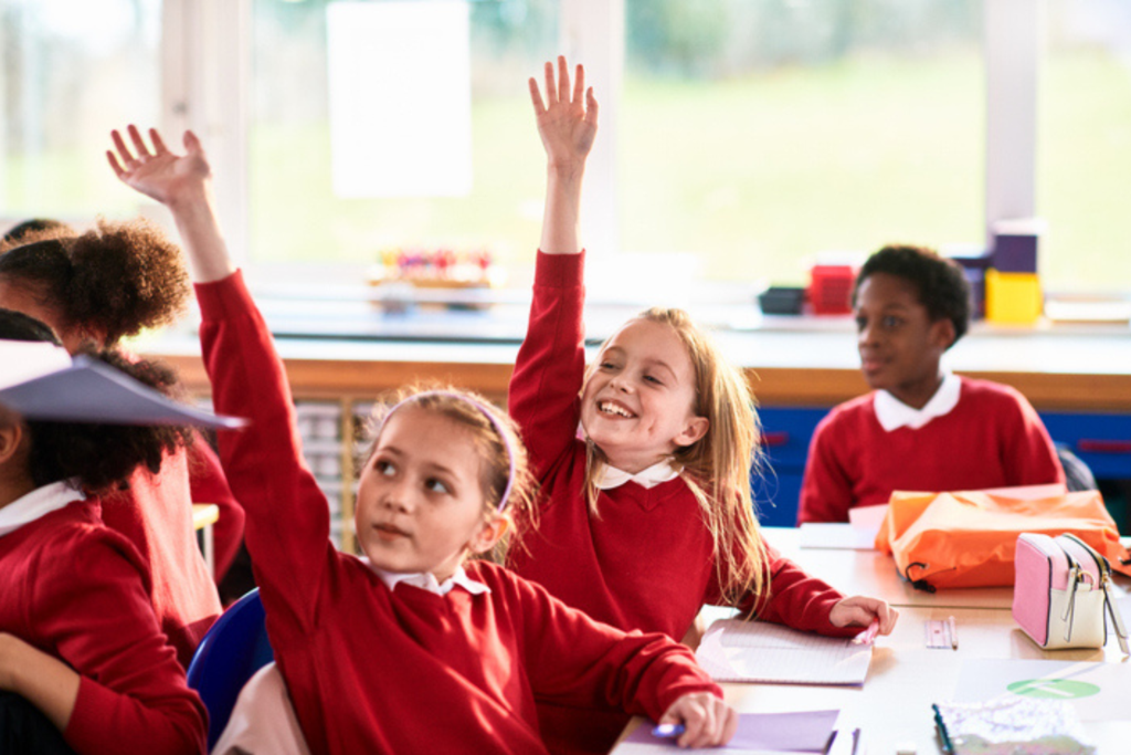 Two smiling schoolgirls in red uniforms raise their hands in a bright classroom.