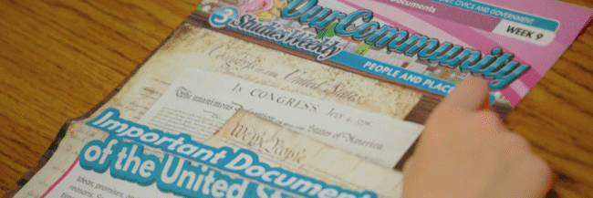 Studies Weekly Our Community week 9 publication on the desk with a student's hand pointing