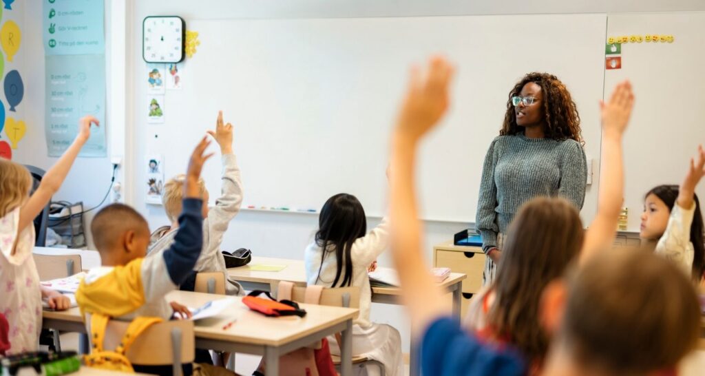 Teacher and students with raised hands Teacher standing at the front of the class, looking at students with their hands raised waiting to answer a question
