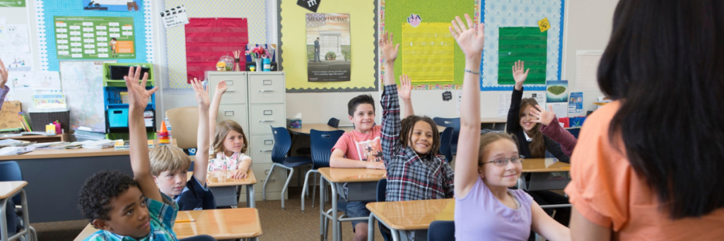 Students in a classroom raising hands to answer a teacher's questions