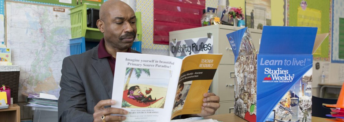 African American teacher at his desk with Studies Weekly products. Studies Weekly blue box